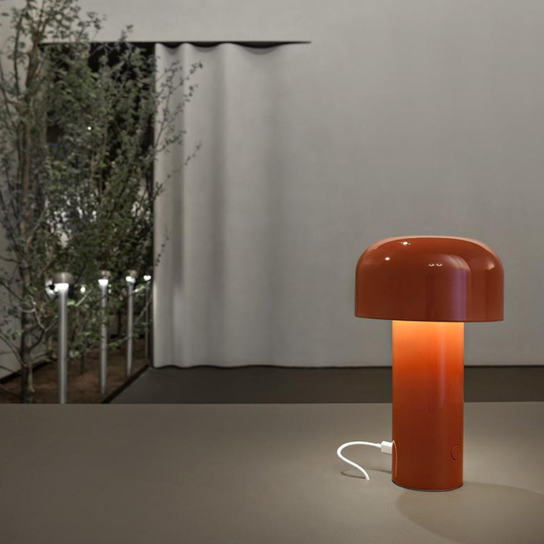Orange mushroom-shaped lamp with a smooth glossy finish, set against a neutral backdrop and subtle green foliage.