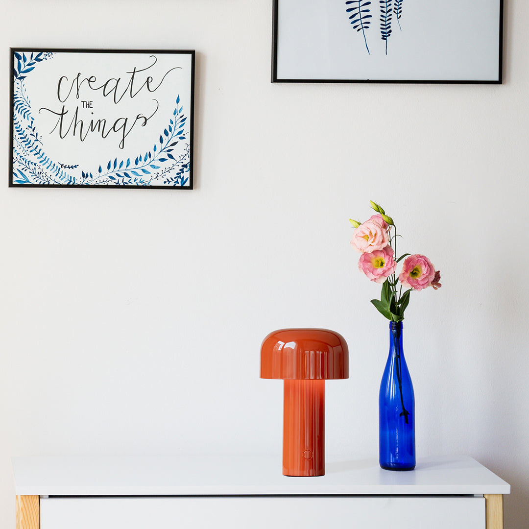"Vibrant orange lamp and blue glass vase with pink flowers against a neutral wall and patterned art."