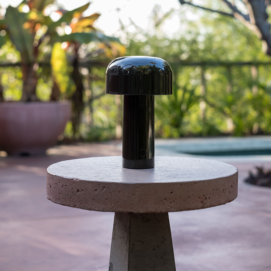 Black mushroom lamp atop a round concrete table in a sunlit outdoor setting, featuring green plants in the background.