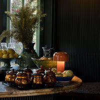Elegant amber jars, green glass bowls, and a warm terracotta lamp on a textured dark table. Rich colors and inviting patterns.