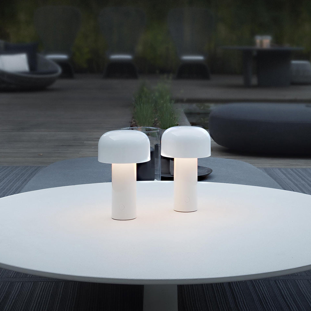 Two sleek white table lamps with a minimalist design, softly illuminated on a round table against a dark outdoor backdrop.
