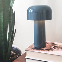 Modern blue lamp with a glossy finish, featuring a cylindrical base and rounded shade. Minimalist design accentuated by plant background.