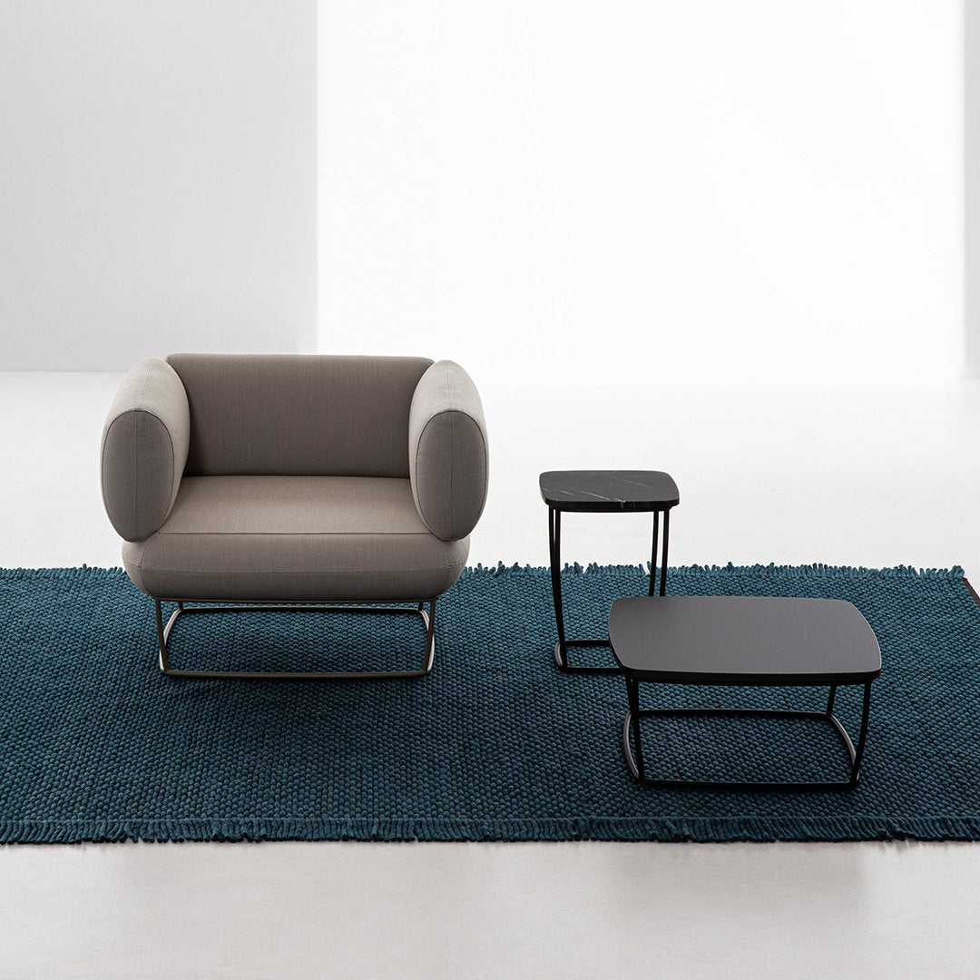 Gray upholstered chair on a dark teal rug with black accent tables, showcasing modern minimalist design.