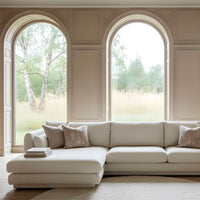 Light beige sectional sofa with subtle texture, accented by soft gray pillows, set against large arched windows.