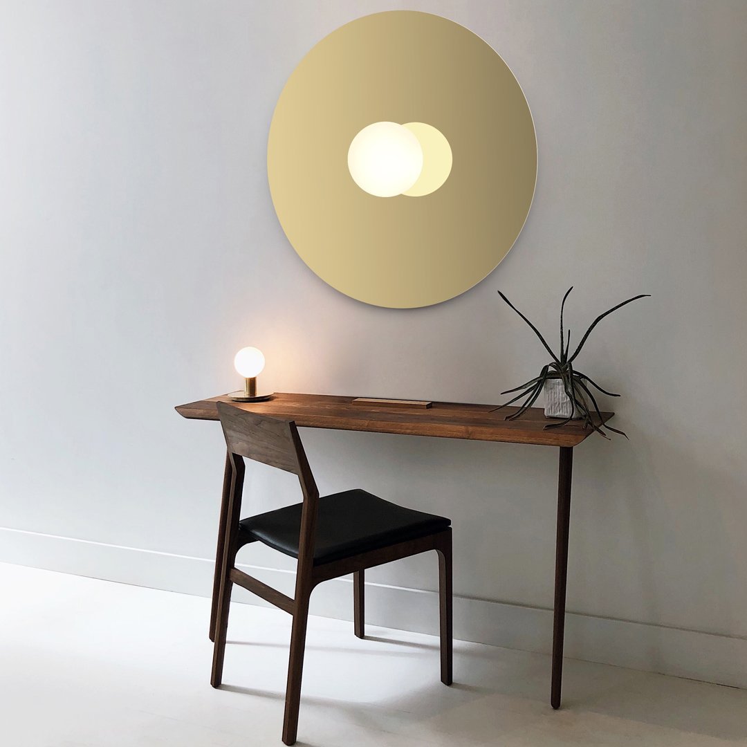 Modern round mirror in soft gold tone above a minimalist wooden desk; sleek black chair and soft white wall backdrop.