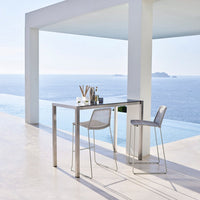 Modern white bar table set with sleek silver accents, featuring transparent chairs against a serene ocean view.