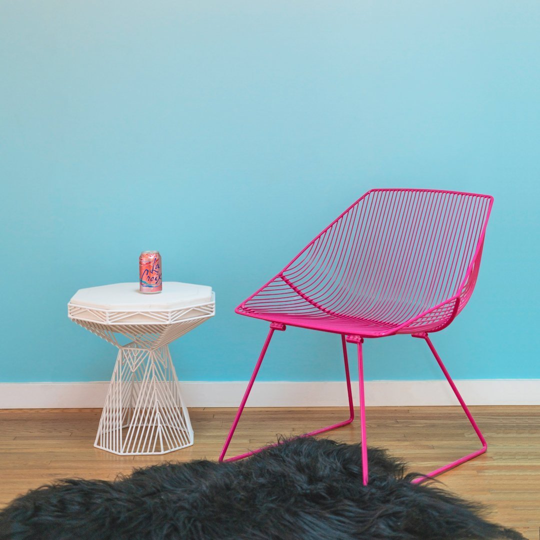 Bright pink wire chair against a soft blue wall, paired with a white geometric side table on a dark textured rug.