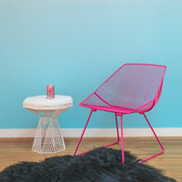 Bright pink wire chair against a soft blue wall, paired with a white geometric side table on a dark textured rug.