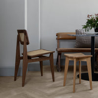 Wooden chair with a natural finish and woven seat, paired with a light wooden stool on herringbone floor pattern.