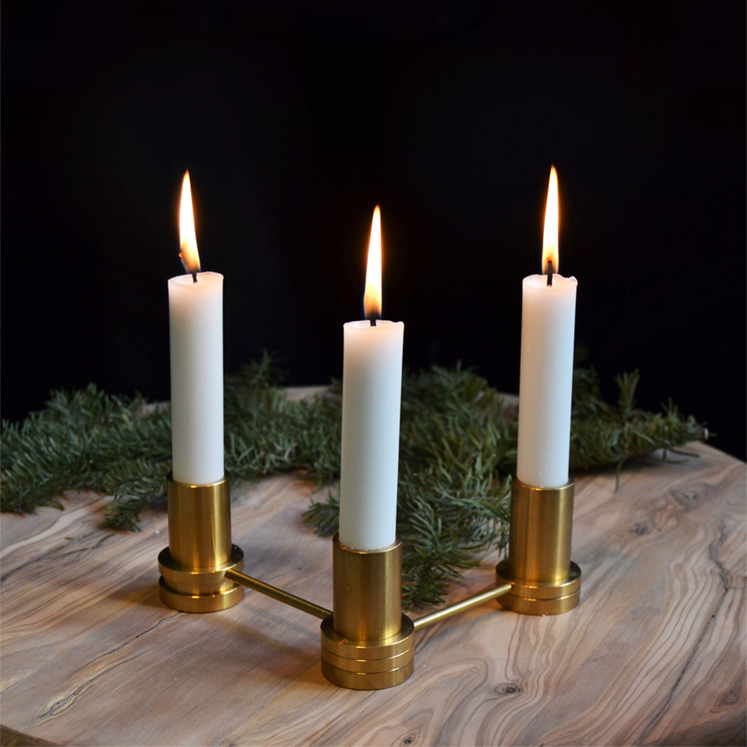 Three white taper candles in a gold candlestick holder on a rustic wooden surface, accented by green foliage.