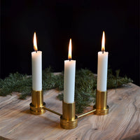 Three white taper candles in a gold candlestick holder on a rustic wooden surface, accented by green foliage.