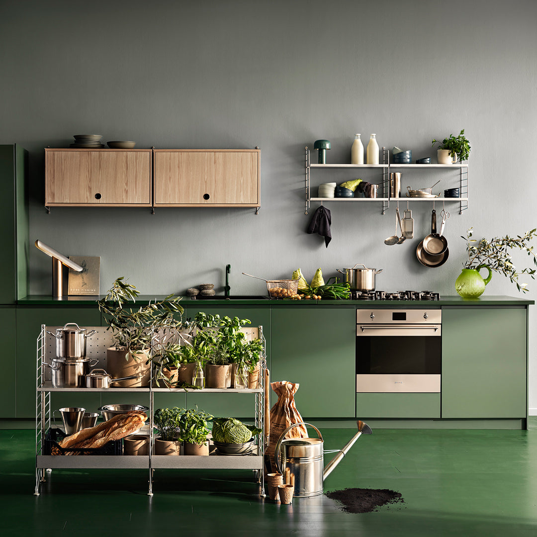 Modern green kitchen with wooden accents, stainless steel shelves, and vibrant plants on display. 