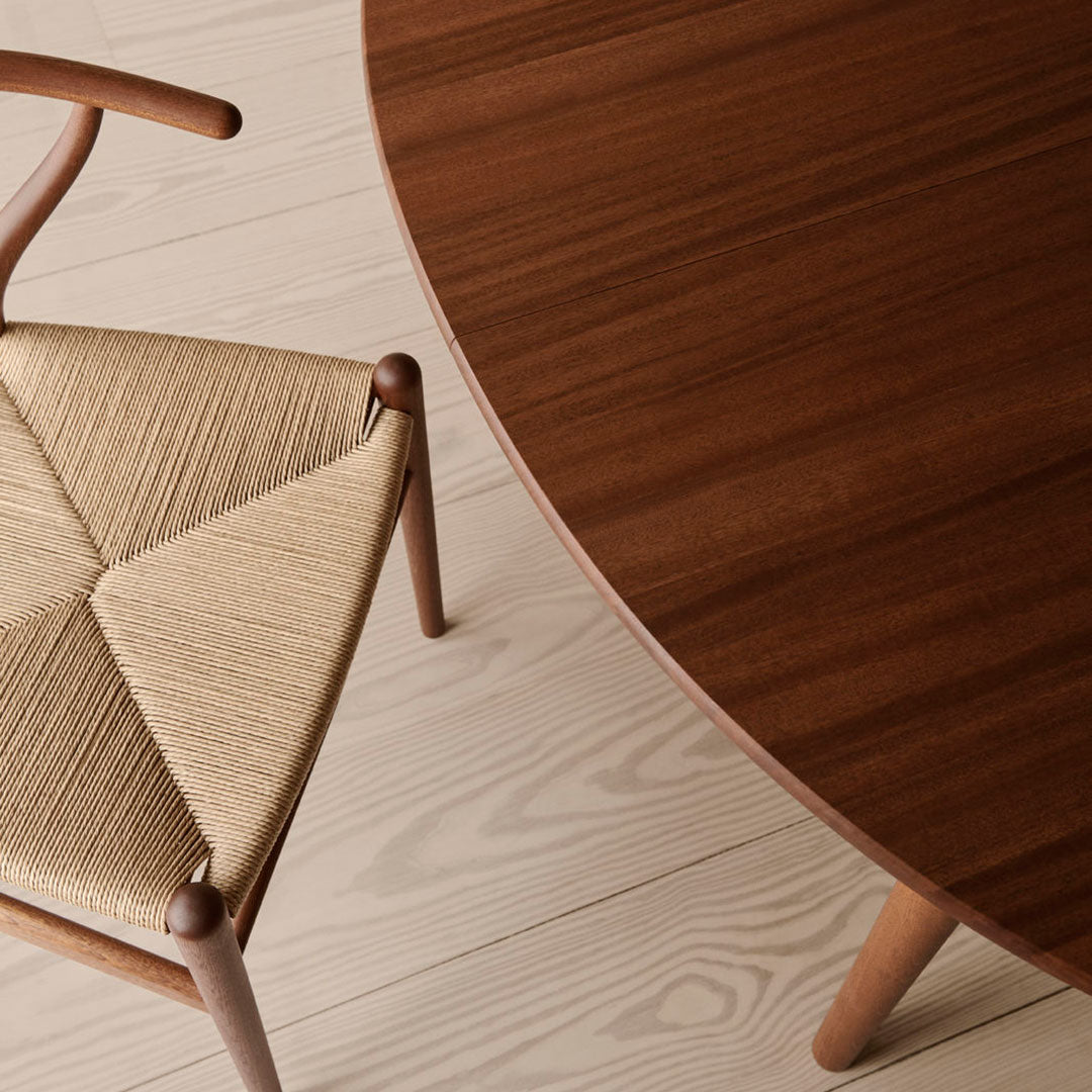 Close-up of a round walnut tabletop and a woven beige chair on light wooden flooring, highlighting rich textures and colors.