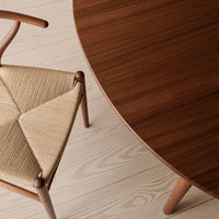 Close-up of a round walnut tabletop and a woven beige chair on light wooden flooring, highlighting rich textures and colors.