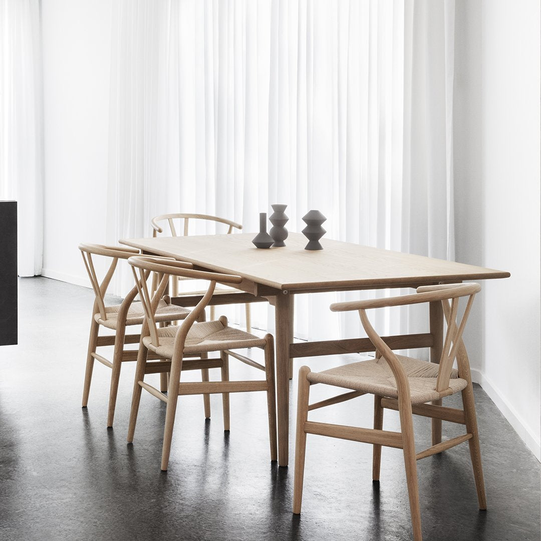 Light wood dining table with natural fiber chairs against a minimalist white backdrop and black floor.