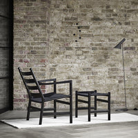 Black wooden chair and stool on a neutral rug, set against a rustic brick wall, creating a modern, minimalist aesthetic.