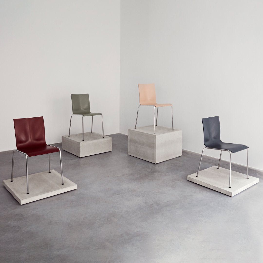 Four modern chairs in burgundy, olive green, peach, and dark gray, displayed on minimalist concrete pedestals.
