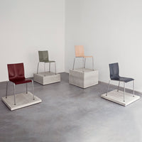 Four modern chairs in burgundy, olive green, peach, and dark gray, displayed on minimalist concrete pedestals.