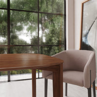 Brown wooden table with a soft beige chair, set against large green-leafed windows and a minimalistic art piece.