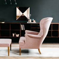 Light pink velvet armchair with gold legs, complemented by a matching ottoman against a dark wall.