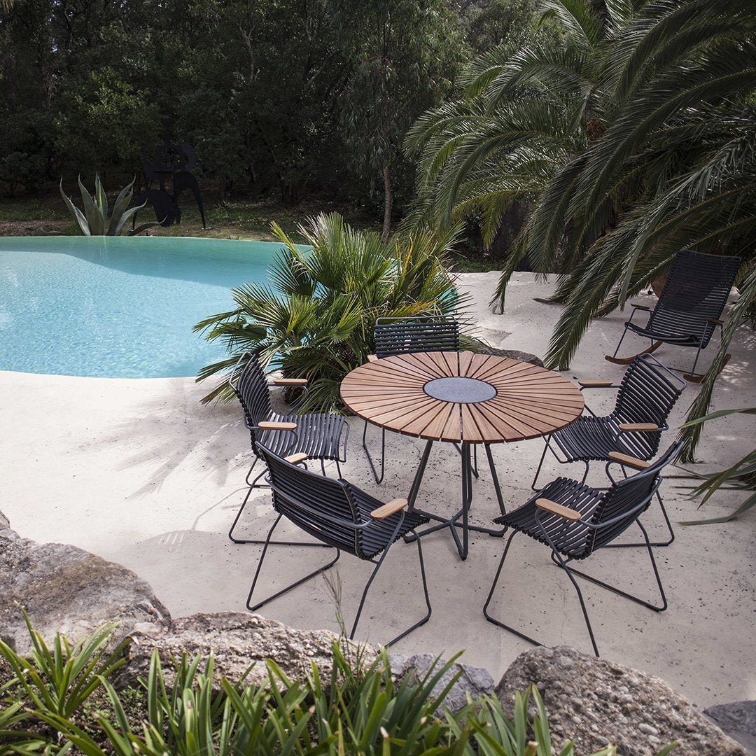 Round wooden table with a sunburst pattern, surrounded by six black mesh chairs, set against lush greenery and a turquoise pool.
