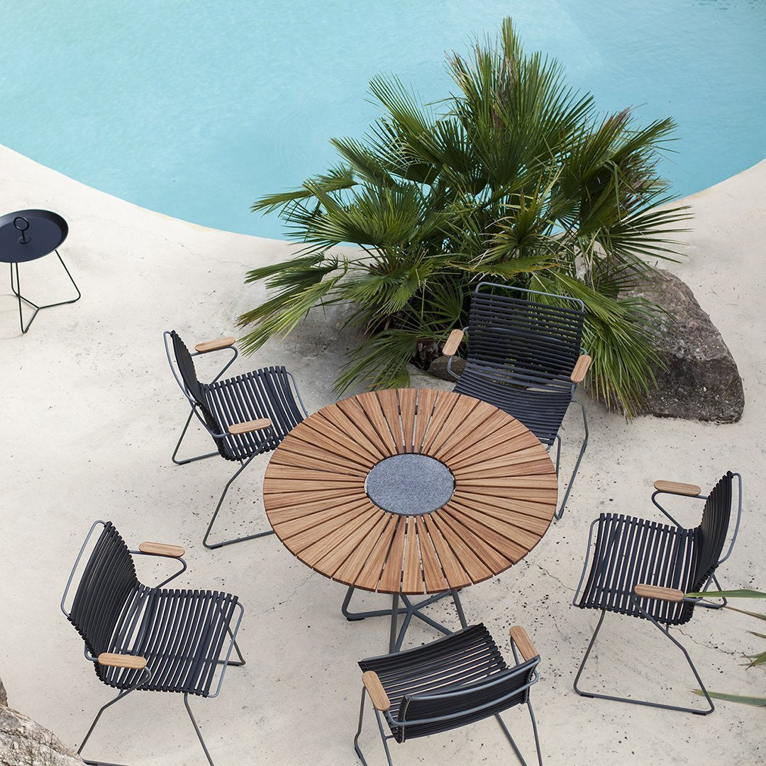 "Black and wooden striped outdoor dining set with circular table and six chairs, surrounded by tropical greenery."