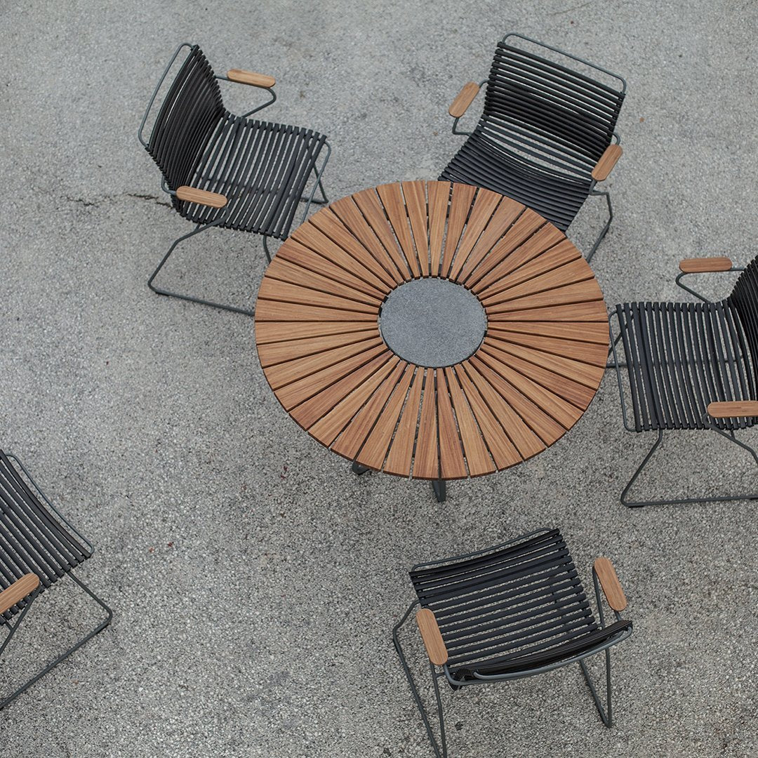 Black metal chairs with wooden armrests surrounding a round wooden slat table with a central circular stone.