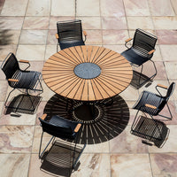 Round wooden table with a central stone, surrounded by six black wire chairs with wooden armrests, on a light stone patio.