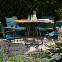 "Round wooden garden table with teal striped chairs, surrounded by green foliage and vibrant flowers."