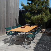 "Natural wood table with green woven chairs, set in a modern outdoor space surrounded by wood and greenery."