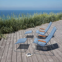 Blue woven patio chairs with wooden armrests on a wooden deck near the ocean, surrounded by lush greenery.