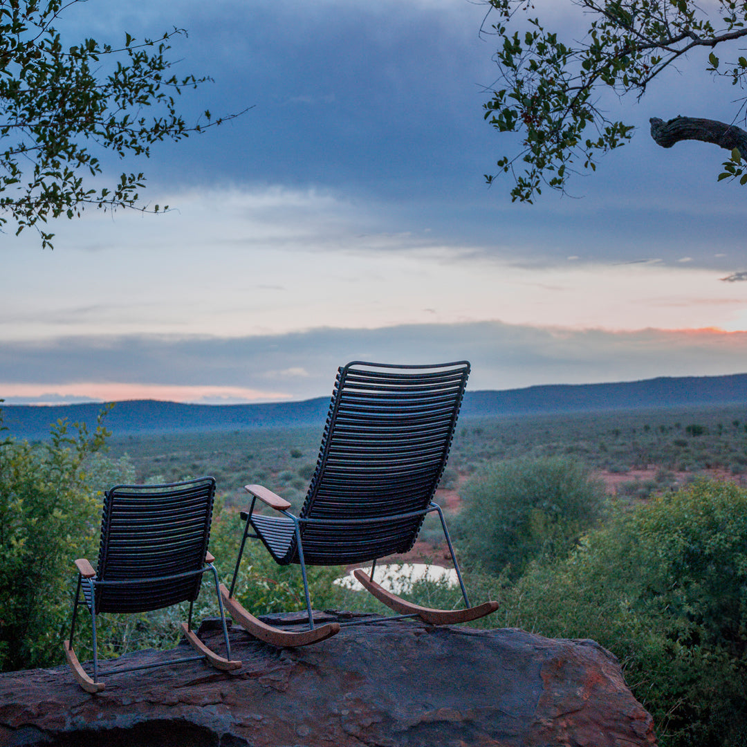 Set of two sleek black rocking chairs against a scenic backdrop of lush green hills and a colorful sunset sky.