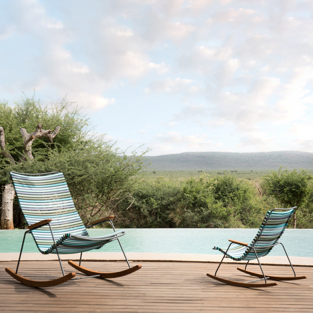"Blue and green striped rocking chairs with wooden frames on a wooden deck, overlooking a serene landscape."