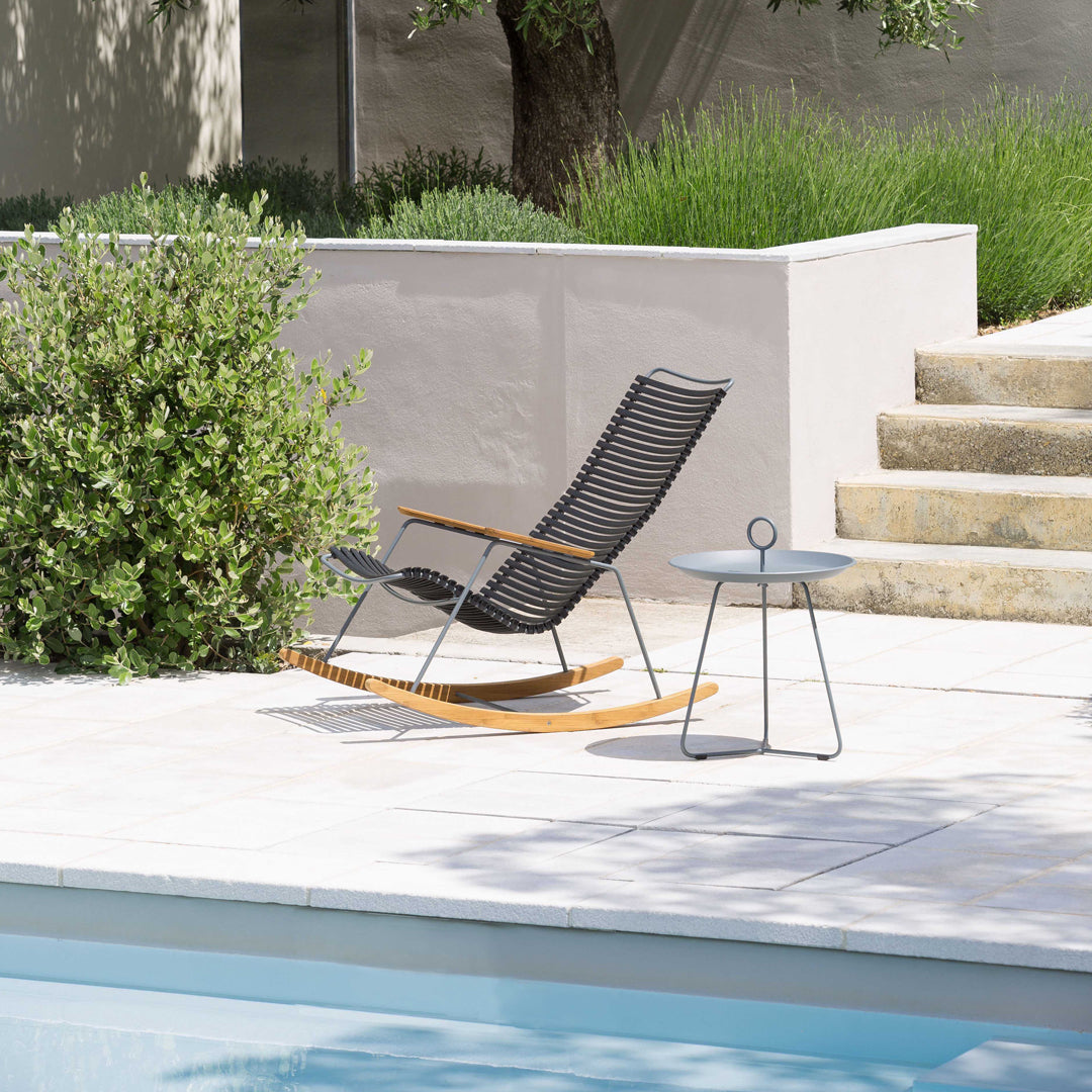 Black striped rocking chair with wooden armrests beside a minimalist round side table in a serene outdoor setting.