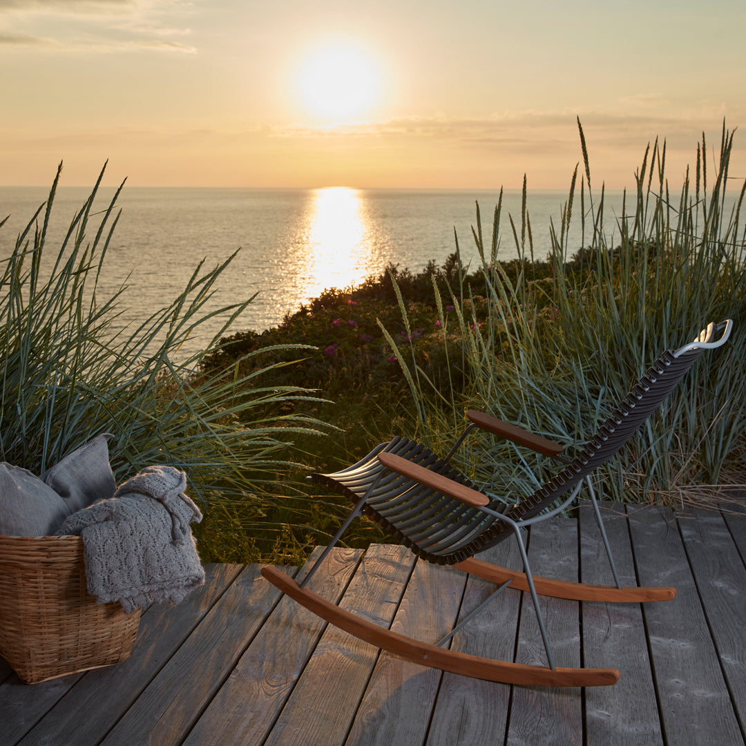 Modern black rocking chair with natural wood accents, set against a sunset backdrop and lush greenery.