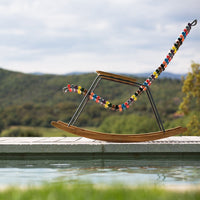 Colorful beaded pattern on a sleek wooden rocking chair with a metallic frame, set against a serene outdoor backdrop.
