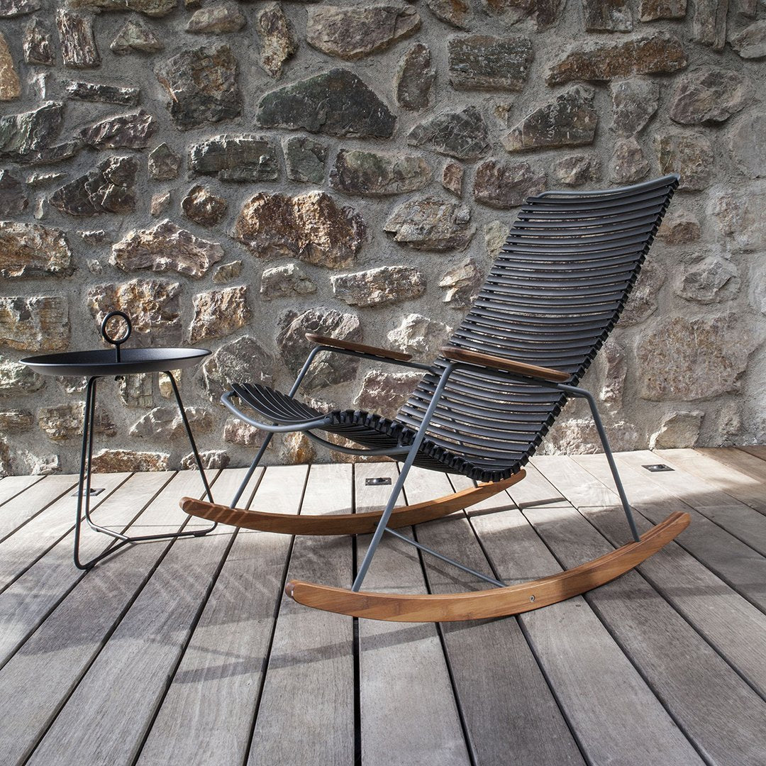 Black woven rocking chair with natural wood accents, paired with a minimalist black side table against a stone wall.