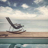Black woven rocking chair on wooden deck with tranquil blue water and cloudy sky backdrop. Modern and stylish design.