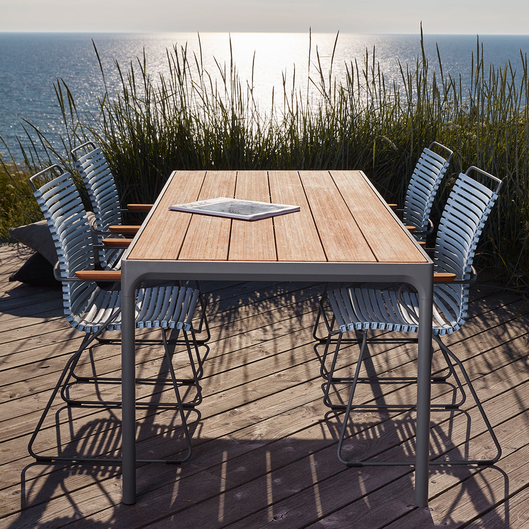 Outdoor dining table with light wood finish and silver metal chairs against a coastal backdrop, featuring natural textures.