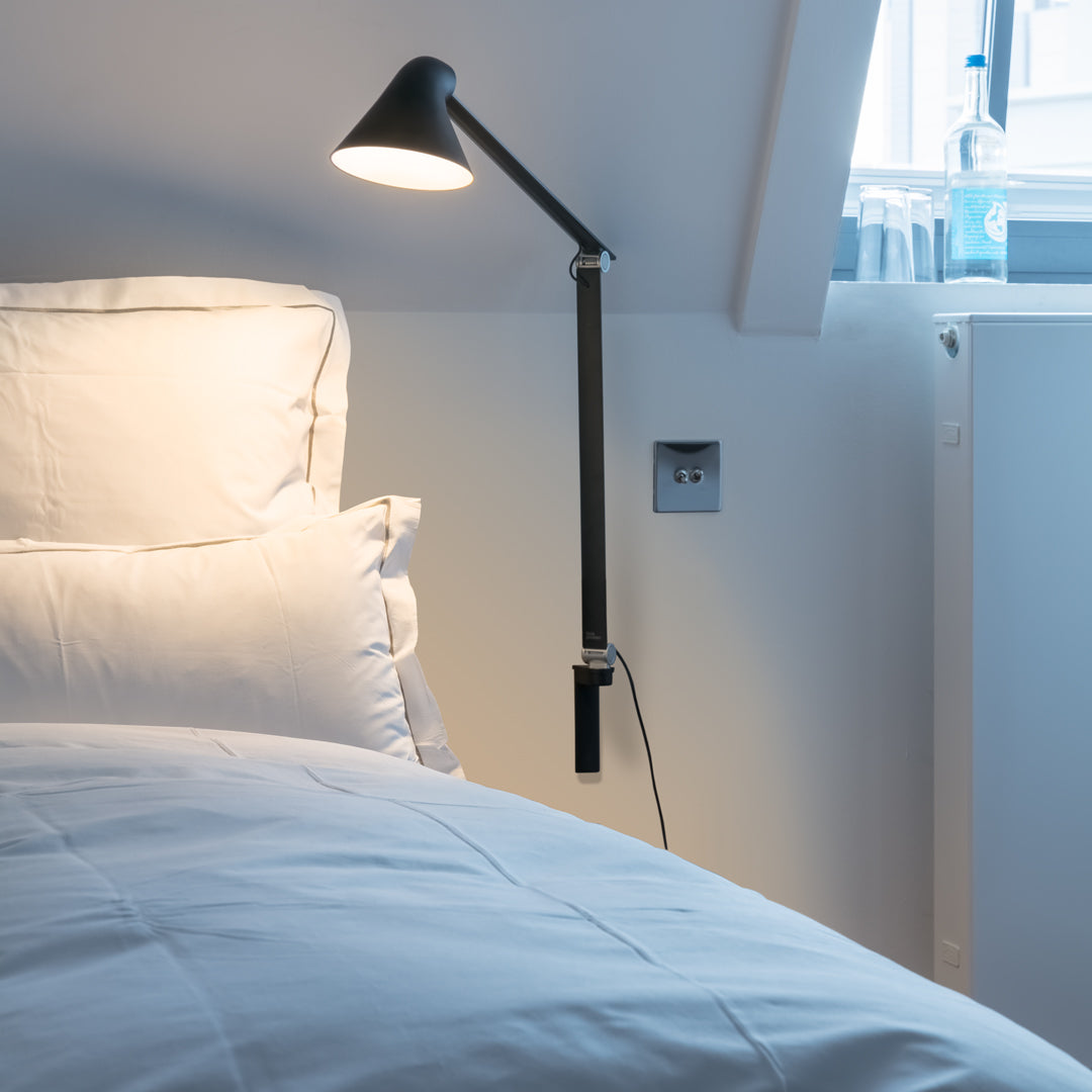 Modern black floor lamp beside a white bed with textured pillows in a bright, minimalistic room.