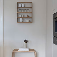 Wooden shelf displaying white and textured glass mugs against a soft beige wall, with a minimalist plant and books below.