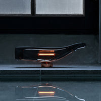 Sleek black glass lamp with warm orange accents, reflecting on a smooth gray surface near a lit window.