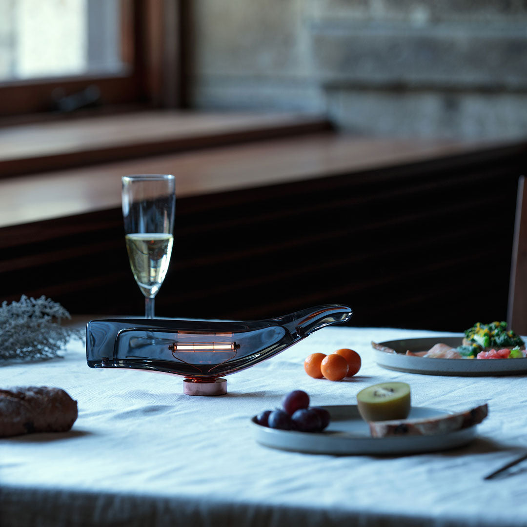Elegant grey glass decanter with copper accents, set on a textured white tablecloth, featuring vibrant fruit and dishes.