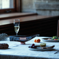 Elegant grey glass decanter with copper accents, set on a textured white tablecloth, featuring vibrant fruit and dishes.