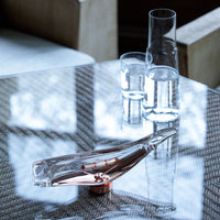 Clear glass water carafe with copper accents, set on a textured gray woven table surface.