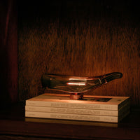 Sleek amber glass lamp with a modern design, illuminated against a rich wooden backdrop and layered neutral books.