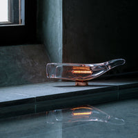 Clear glass lamp with warm amber accents, featuring sleek, modern design on a gray stone surface.