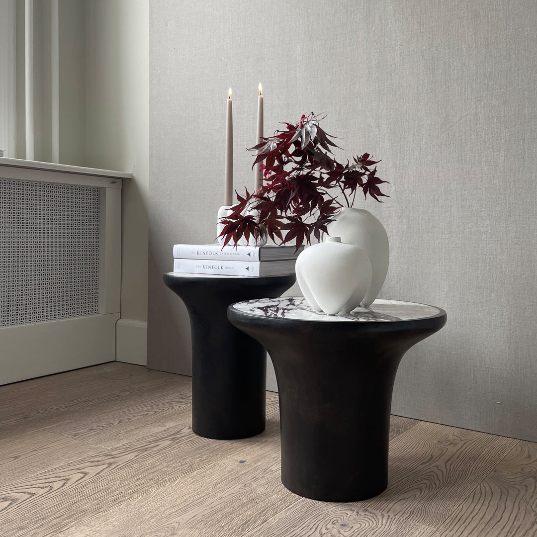 "Black and white accent tables with smooth curves, complemented by a minimalist vase and rich maroon foliage."