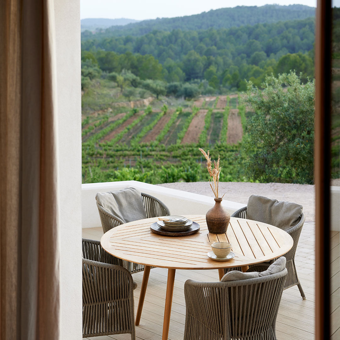 "Natural wood round table with grey woven chairs, set against a vibrant green vineyard backdrop."