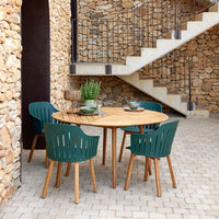 Round wooden dining table with teal chairs and a natural stone backdrop, featuring earthy tones and a contemporary design.